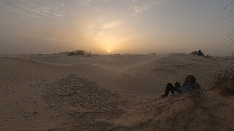 Un trekkeur assis sur une dune, contemplant un somptueux coucher de soleil dans le désert tunisien, plongé dans une atmosphère paisible et immersive.